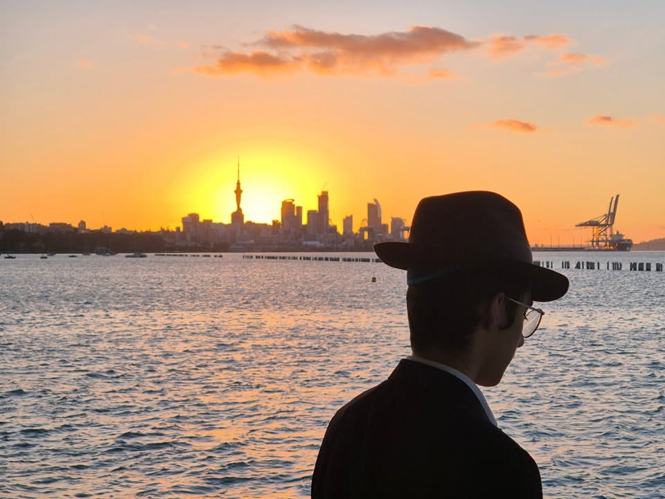 A man in a hat looking across Auckland harbour towards the city skyline at sunset