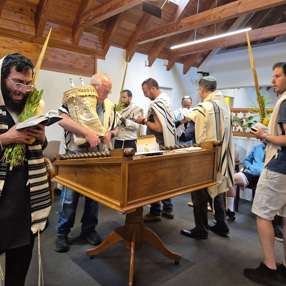 Men in tallitot holding lulav and etrog around a wooden bimah with a Torah scroll during Sukkot