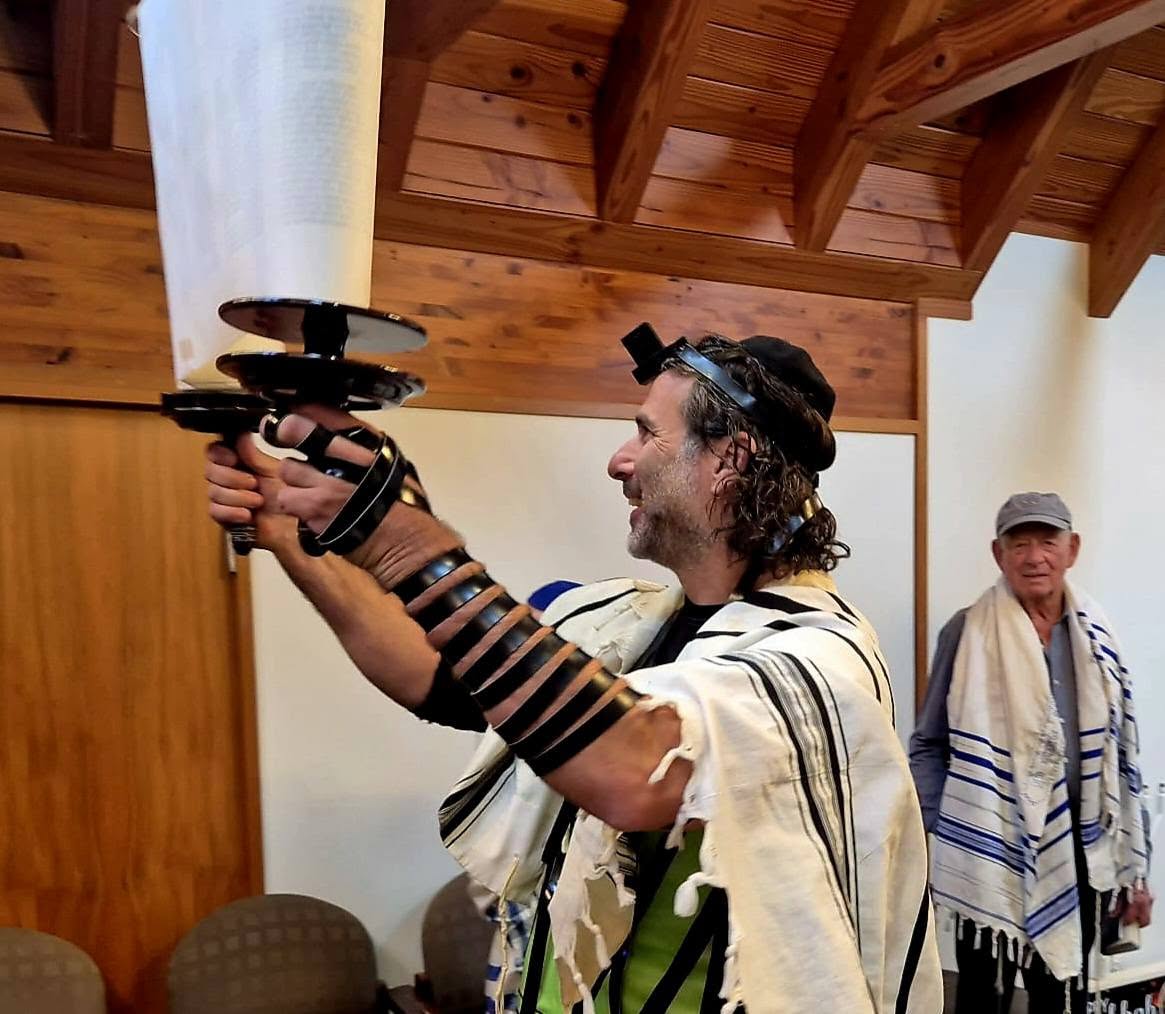 A man in tallit and tefillin lifting an open Torah scroll beneath the timber ceiling at 788