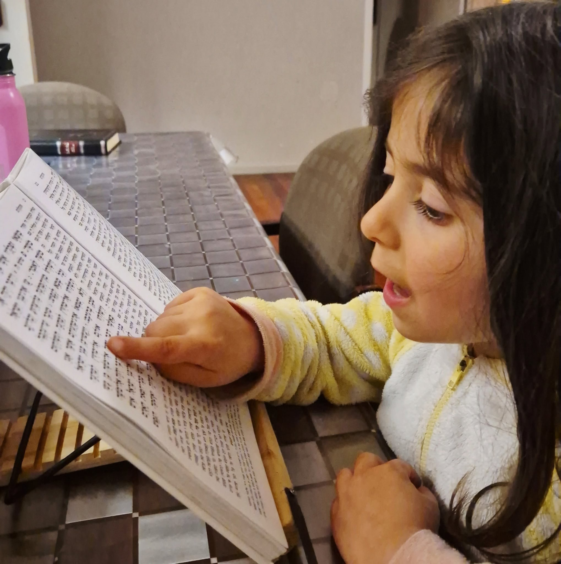 A young girl pointing to Hebrew text in an open prayer book and reading aloud