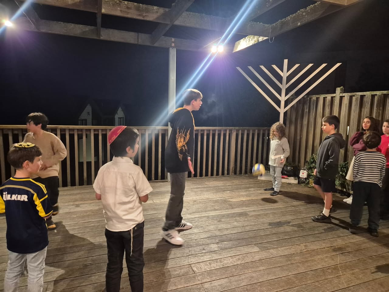 Children playing on a wooden deck at night beside a large wooden menorah