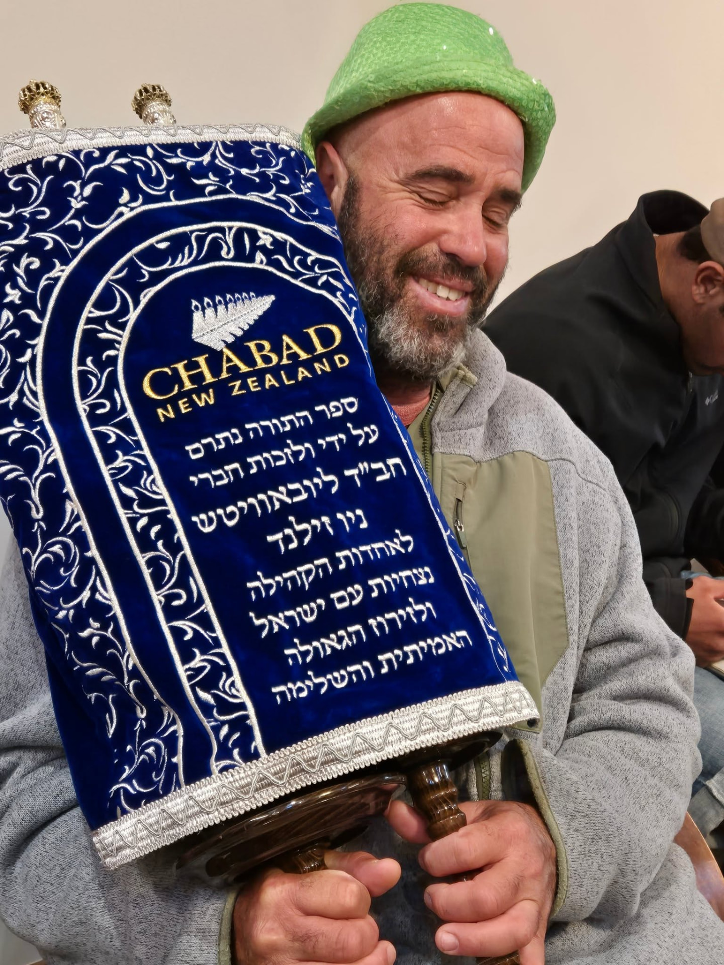 A smiling man holding a Torah scroll in a blue velvet mantle embroidered Chabad New Zealand