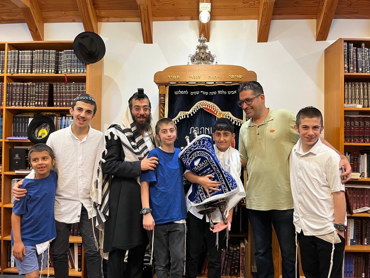 Boys and adults standing with a Torah scroll in front of the ark, surrounded by bookshelves