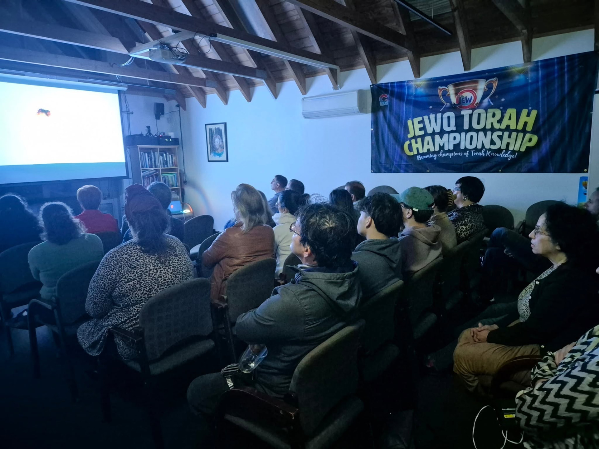 A community audience seated watching a projected screening at 788 beneath a JewQ Torah Championship banner
