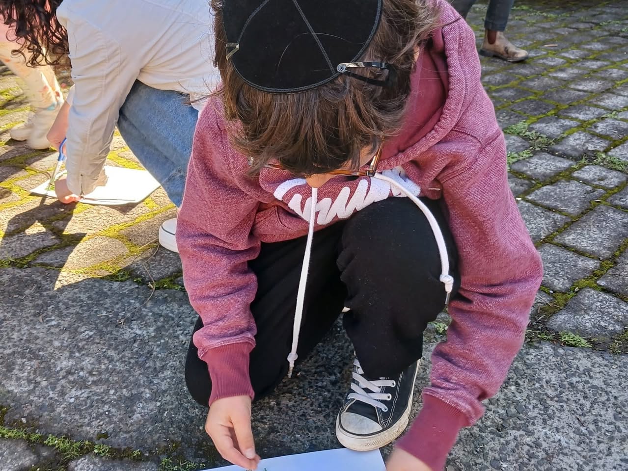 A child in a kippah crouching on paving stones, drawing on a sheet of paper
