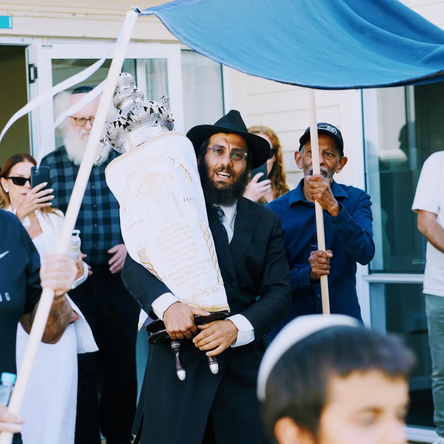 A rabbi carrying a Torah scroll beneath a blue canopy outside the building, surrounded by community members