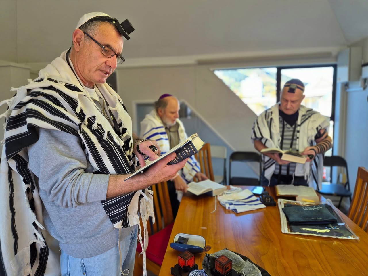 Men in tallit and tefillin reading from prayer books around a wooden table at 788